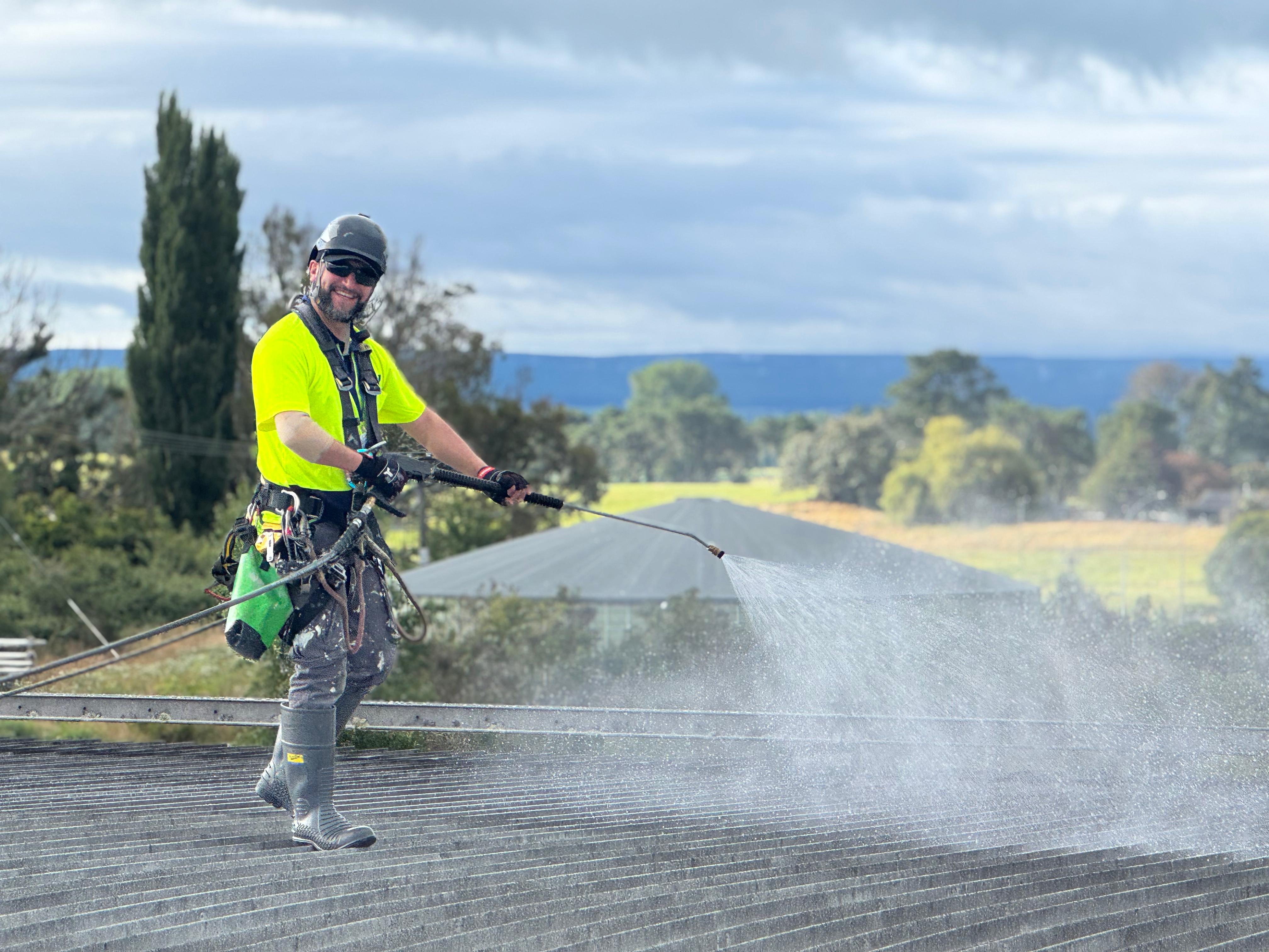 Helmeted crew member in yellow high-vis clothing, gloves and a harness washing a roof