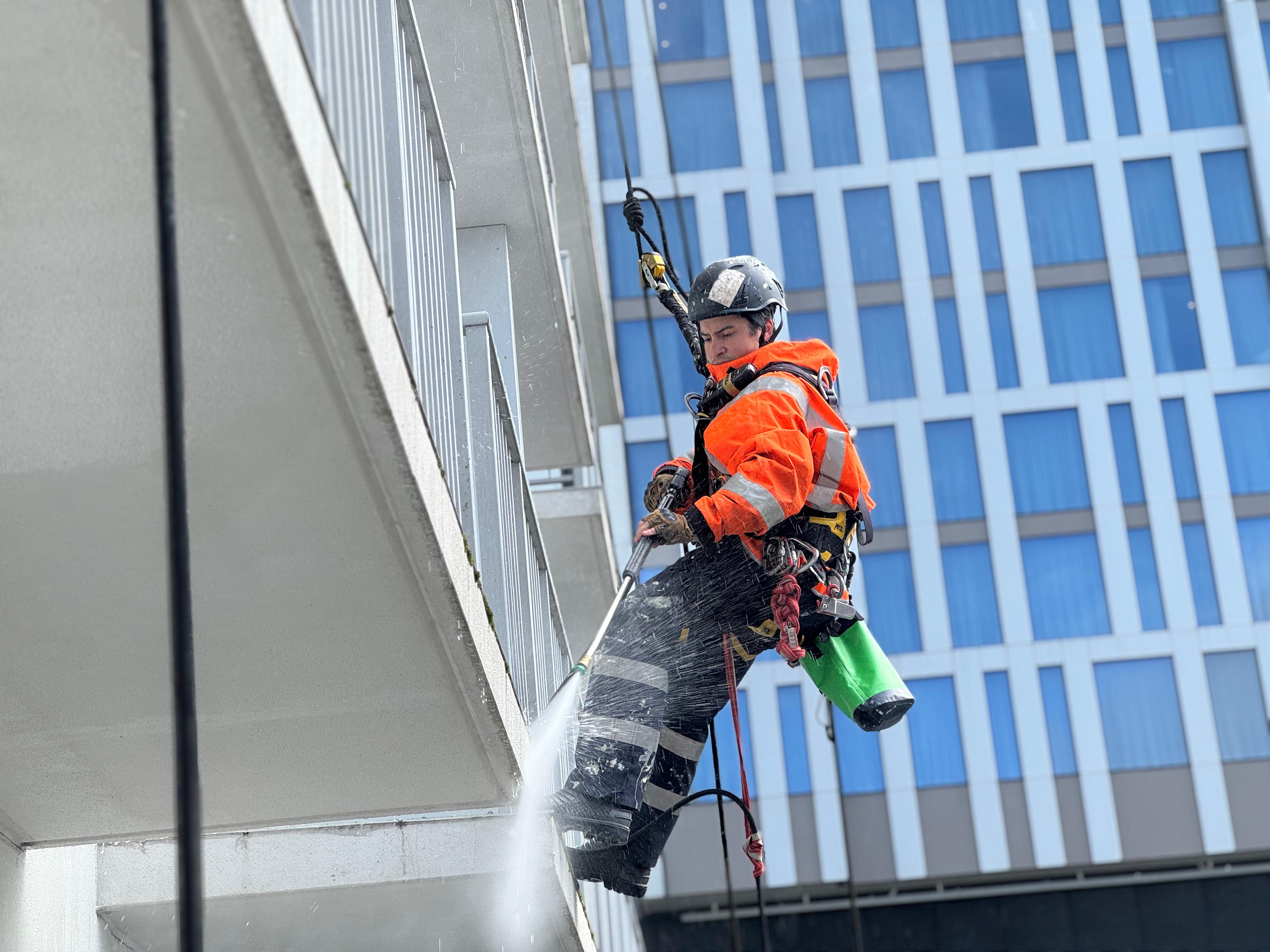 Helmeted rope-access technician in orange high-vis clothing washing a building facade