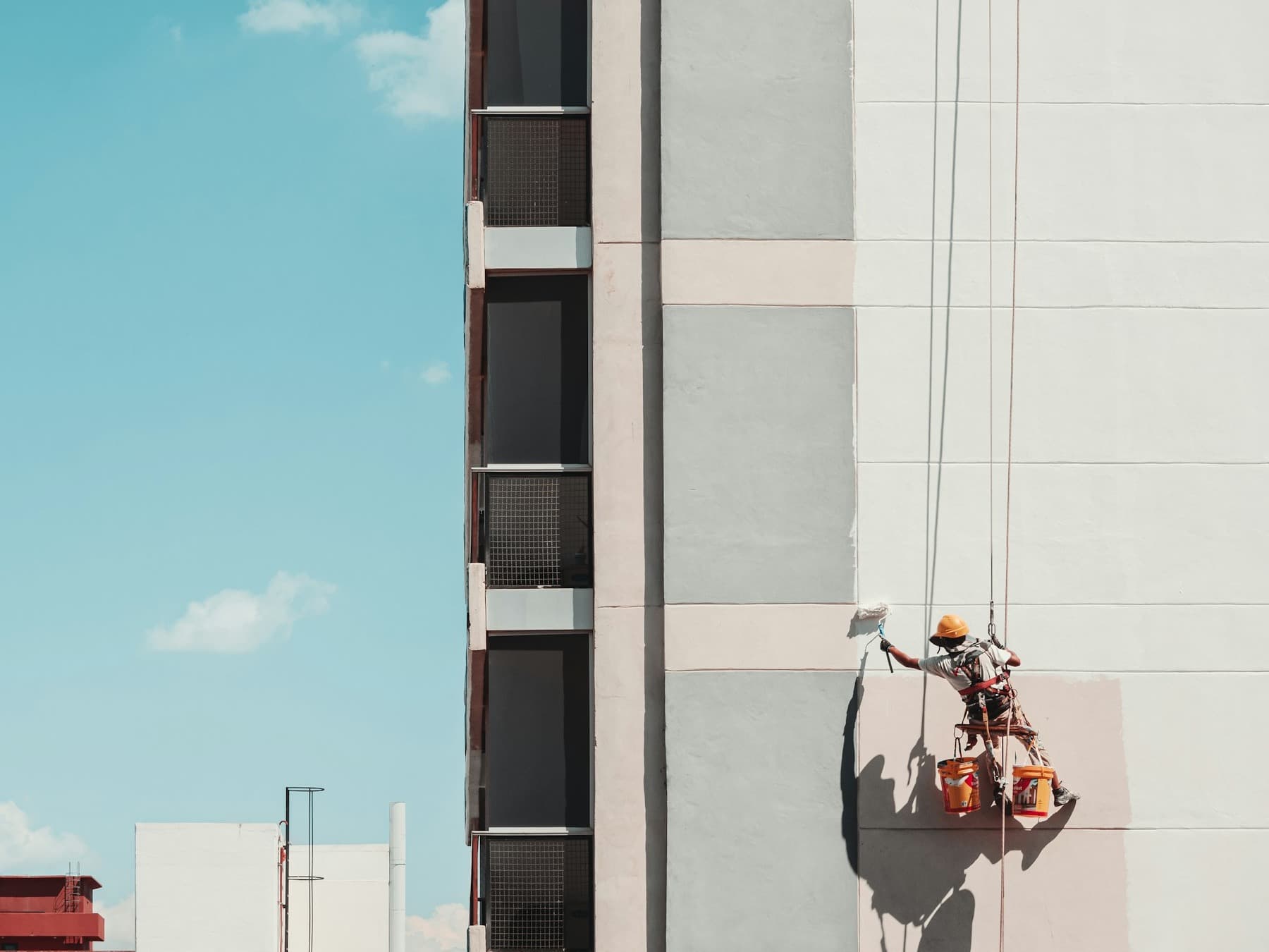 A rope access painter in an orange helmet rolling a fresh coat onto a pale rendered facade under a blue sky, paint tins clipped below him.