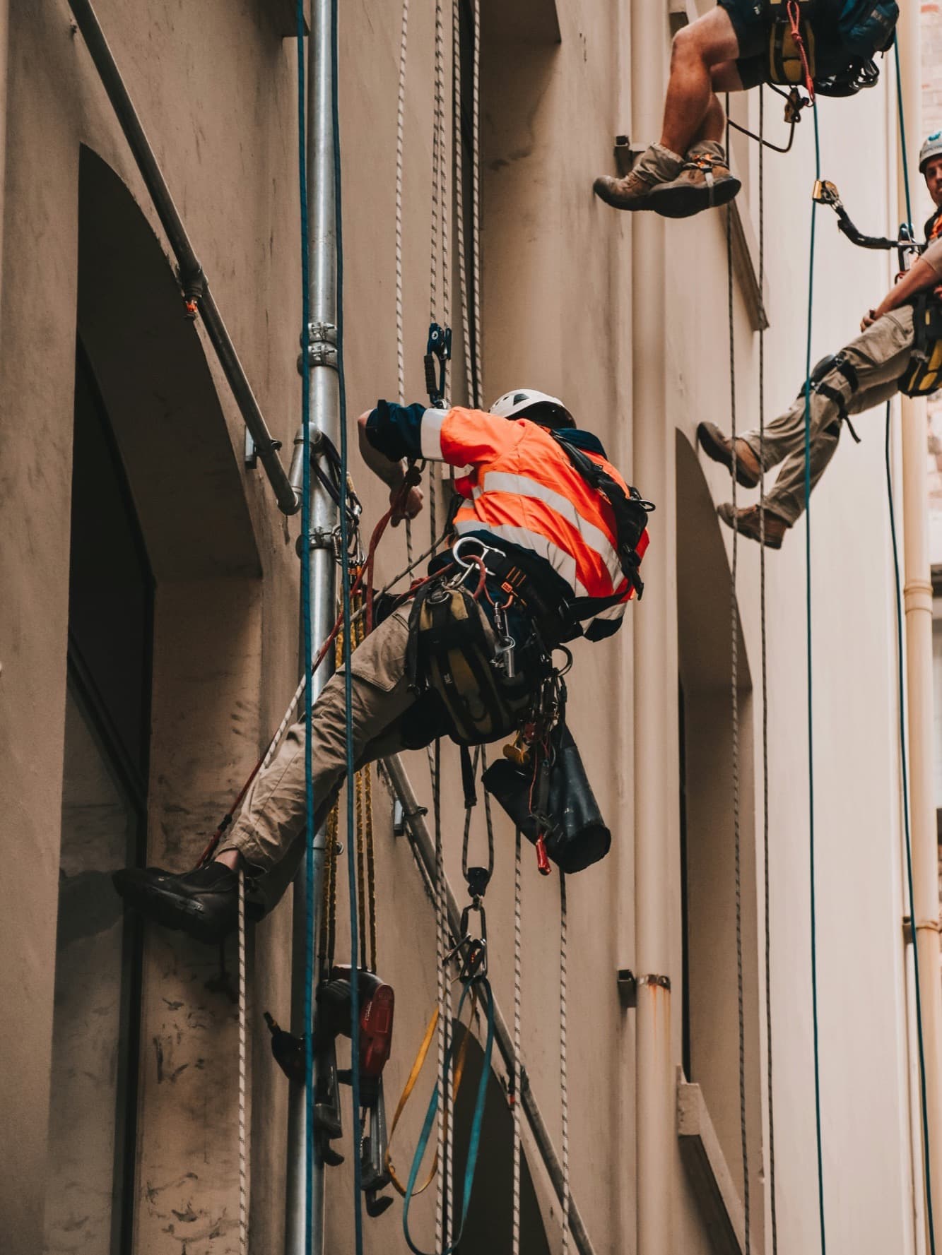 A rope access technician in orange hi-vis and a white helmet on rope against a rendered facade, tools clipped to the harness.