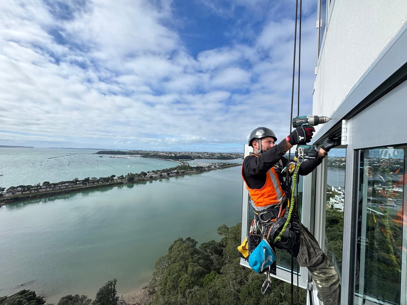 A rope access technician on ropes, drilling a window frame high above the water, the Auckland coastline stretching behind him.