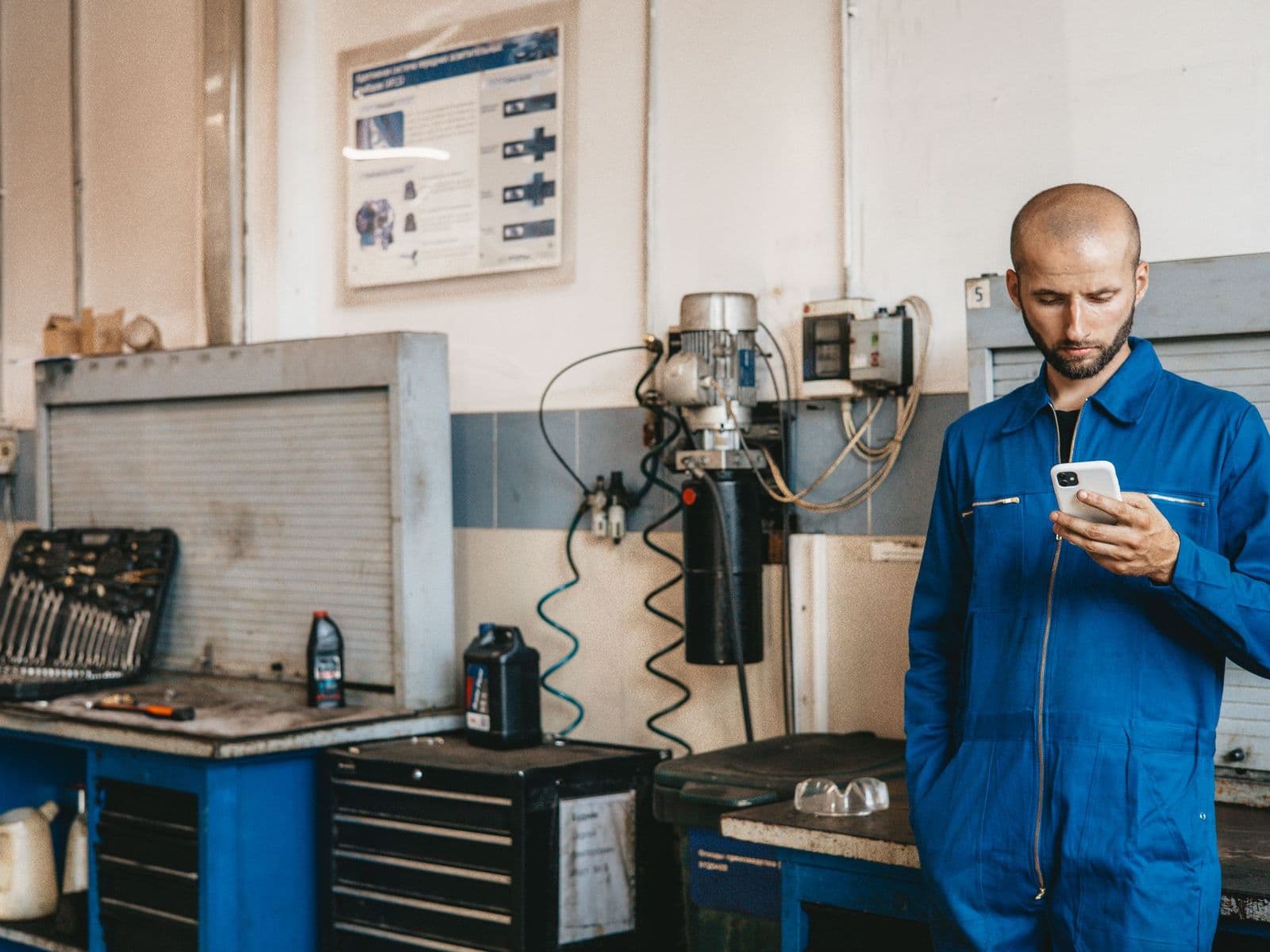 A tradesperson in blue overalls checking his phone in a workshop, beside a workbench, a tool chest and an open spanner set.